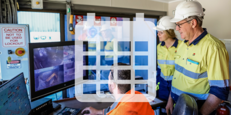 A mine worker is working on a computer with several screens, whilst two inspectors look over his shoulder.