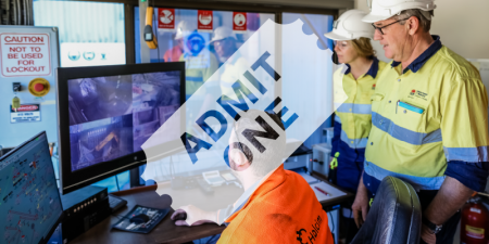 Photograph of two people standing behind a man sitting at a desk. On the desk there are several screens showing machinery working on a mine site. There is a ticket icon laid over the image that reads 'admit one'.