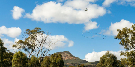A helicopter flying across a blue sky. Trees and a small mountain in the bottom third of the image.